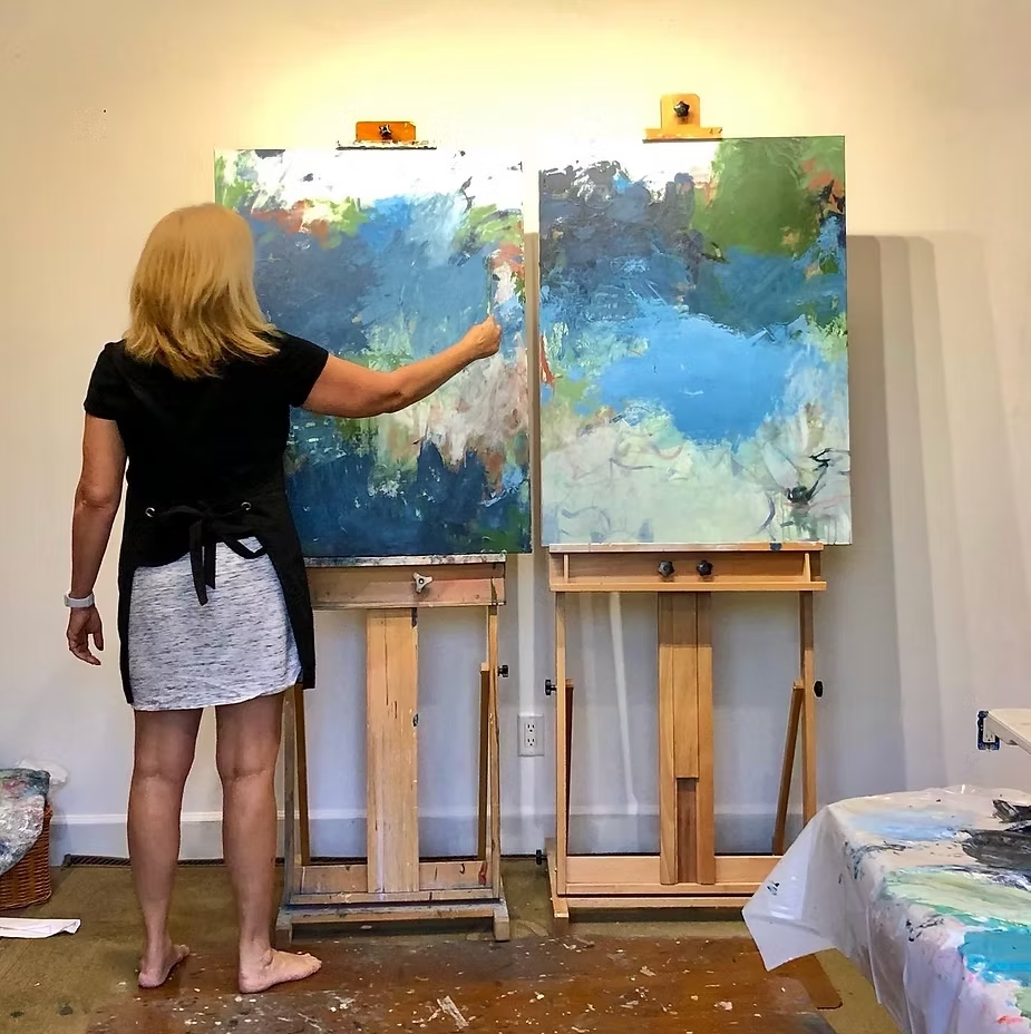 Woman painting on two canvases in art studio. She wears black top and white skirt. Paintings are abstract with blue and green hues.