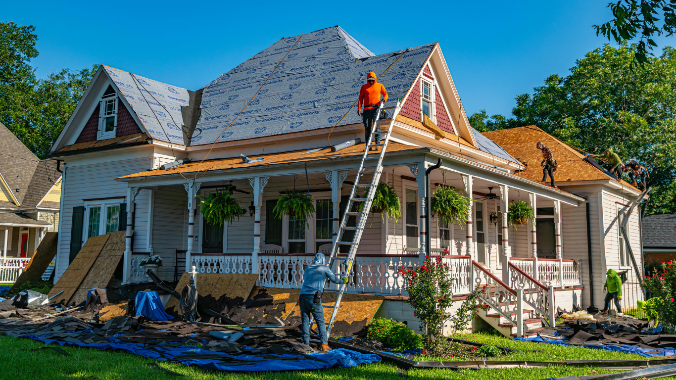 Workers reroofing a white house with gray shingles, one on a ladder and one on the roof