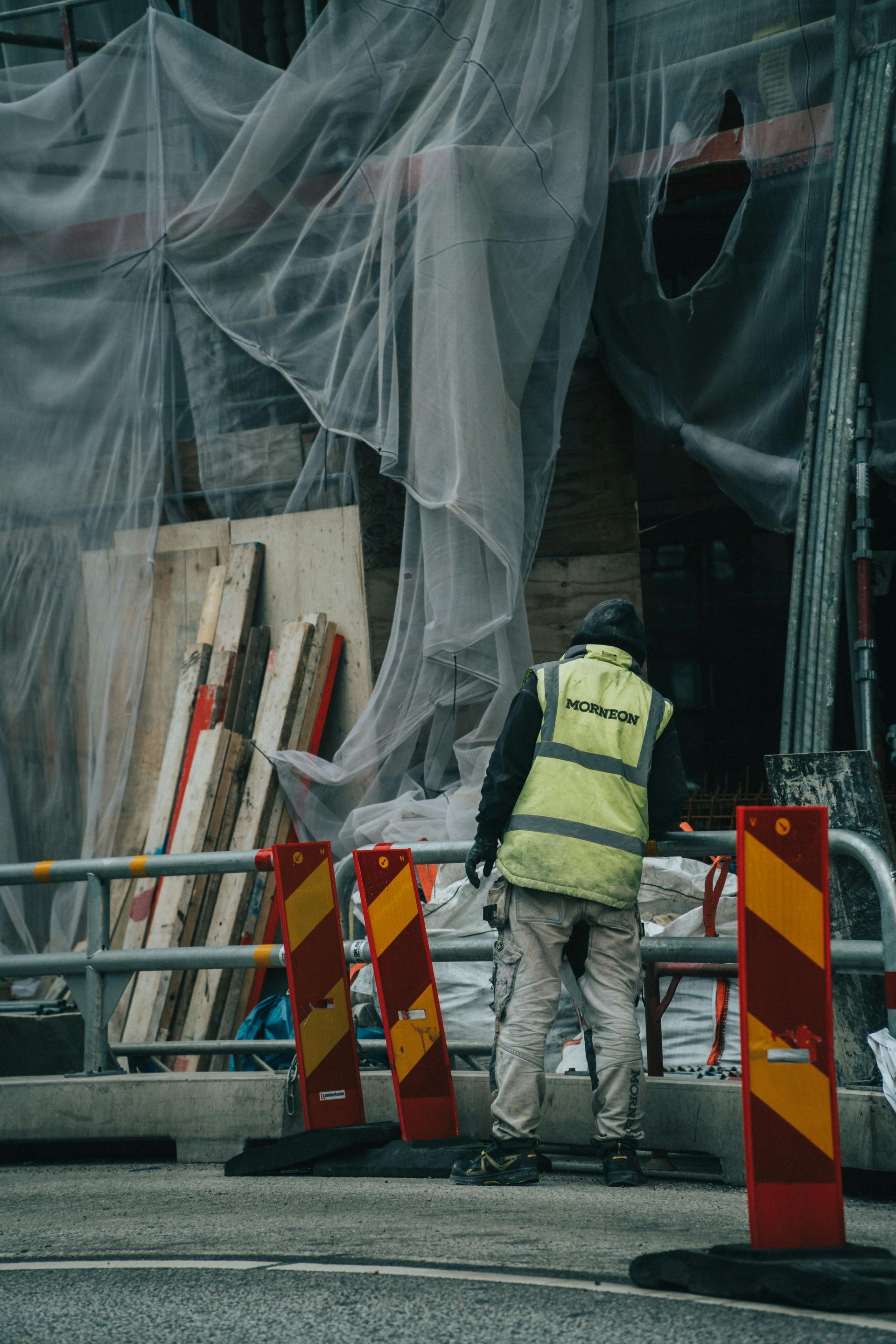 Construction worker in a yellow vest standing by a netted building facade with red road barriers.