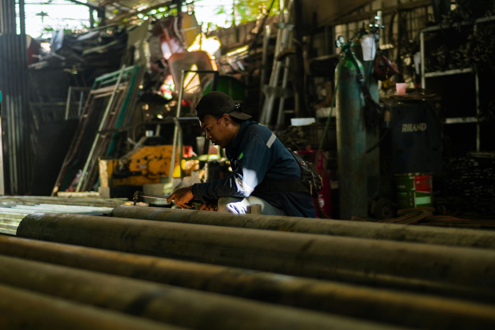 Worker welding metal pipes in a dim industrial workshop, sparks visible.