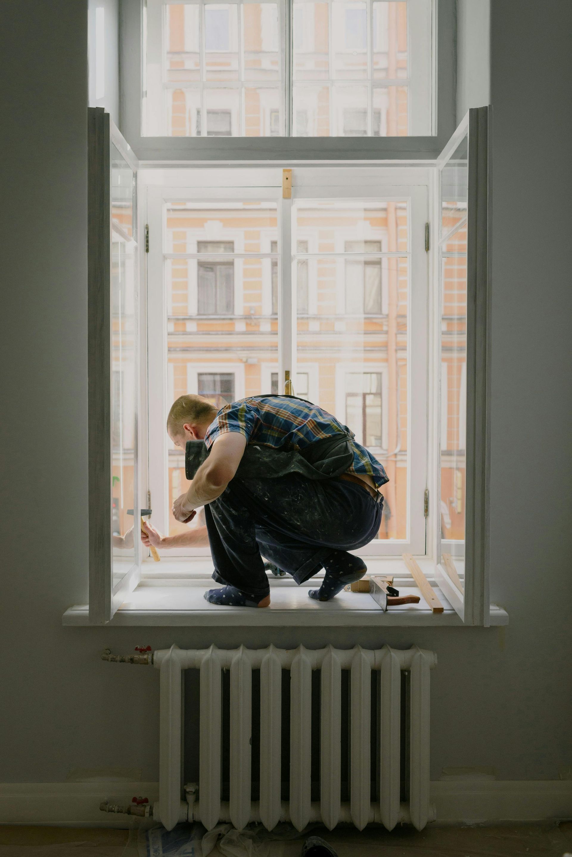 Person crouching on a windowsill inside a room, looking out at a building across the street.