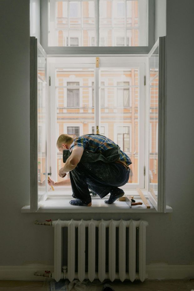 Person crouching on a window ledge inside a room, looking out at buildings across the street.
