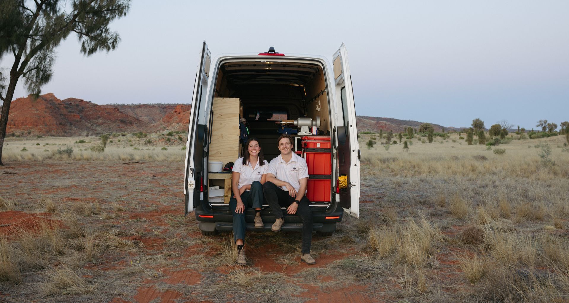 Two people sit on the back of a van in a desert landscape.