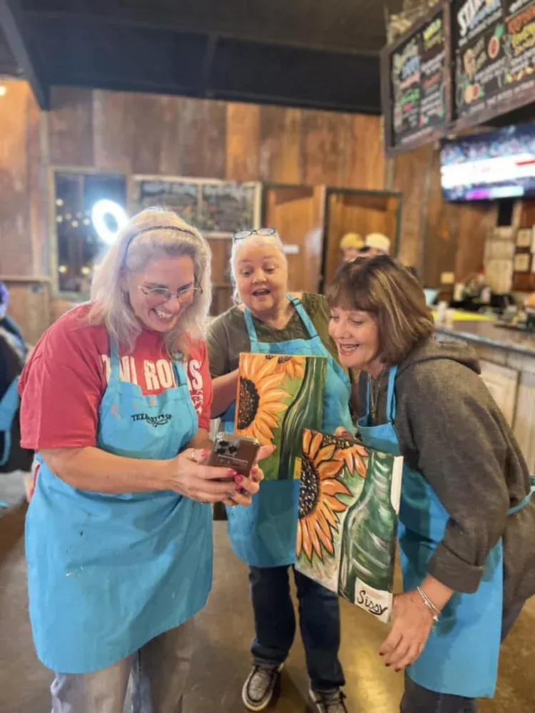 Three smiling staff members in aprons huddle around a phone in a restaurant counter area.