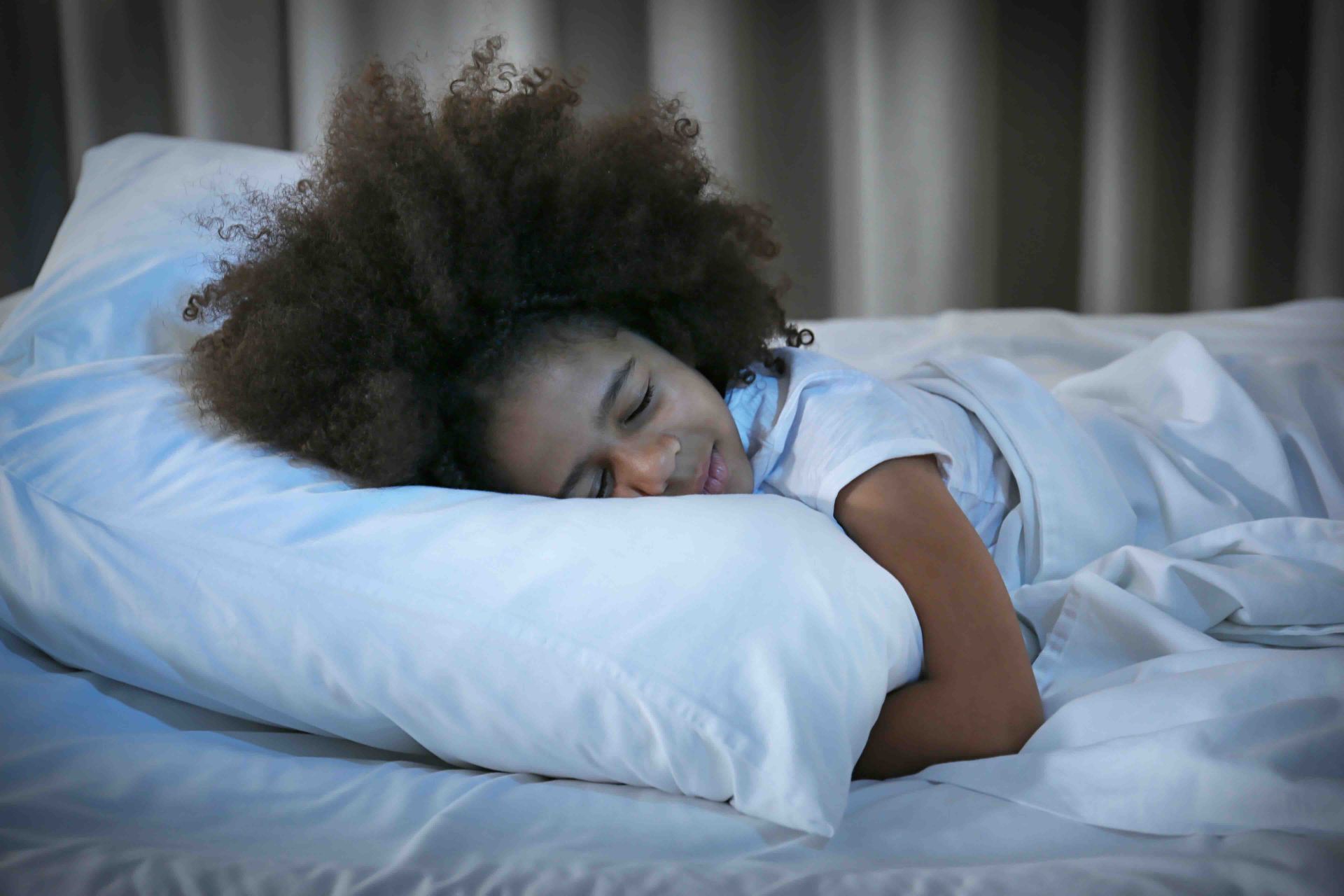 Person sleeping on a bed with a white pillow and white sheets in a dim room