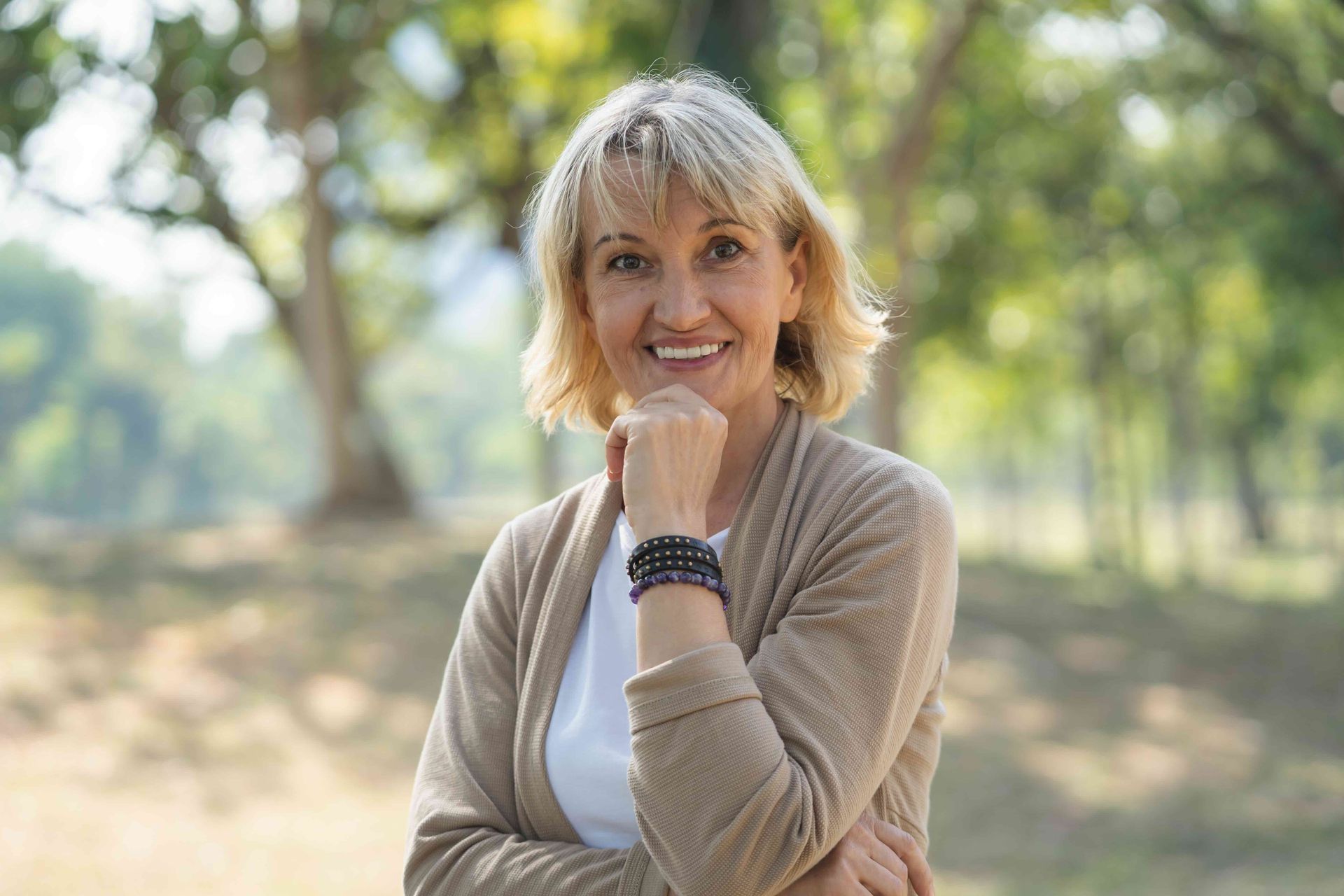 Smiling woman in a beige cardigan standing outdoors with arms crossed in a sunny park