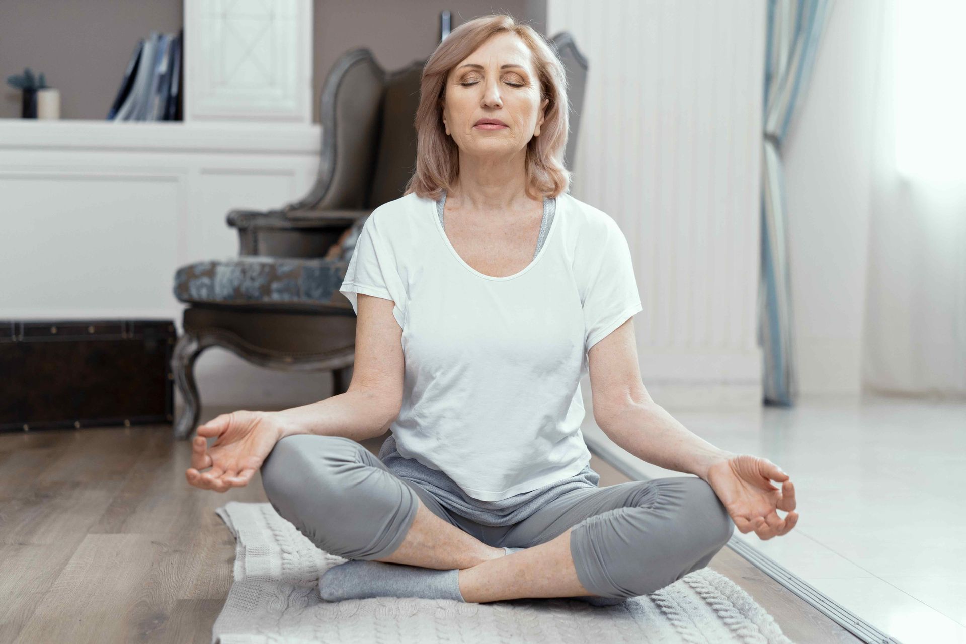 Woman sitting cross-legged on a yoga mat, meditating in a bright living room.