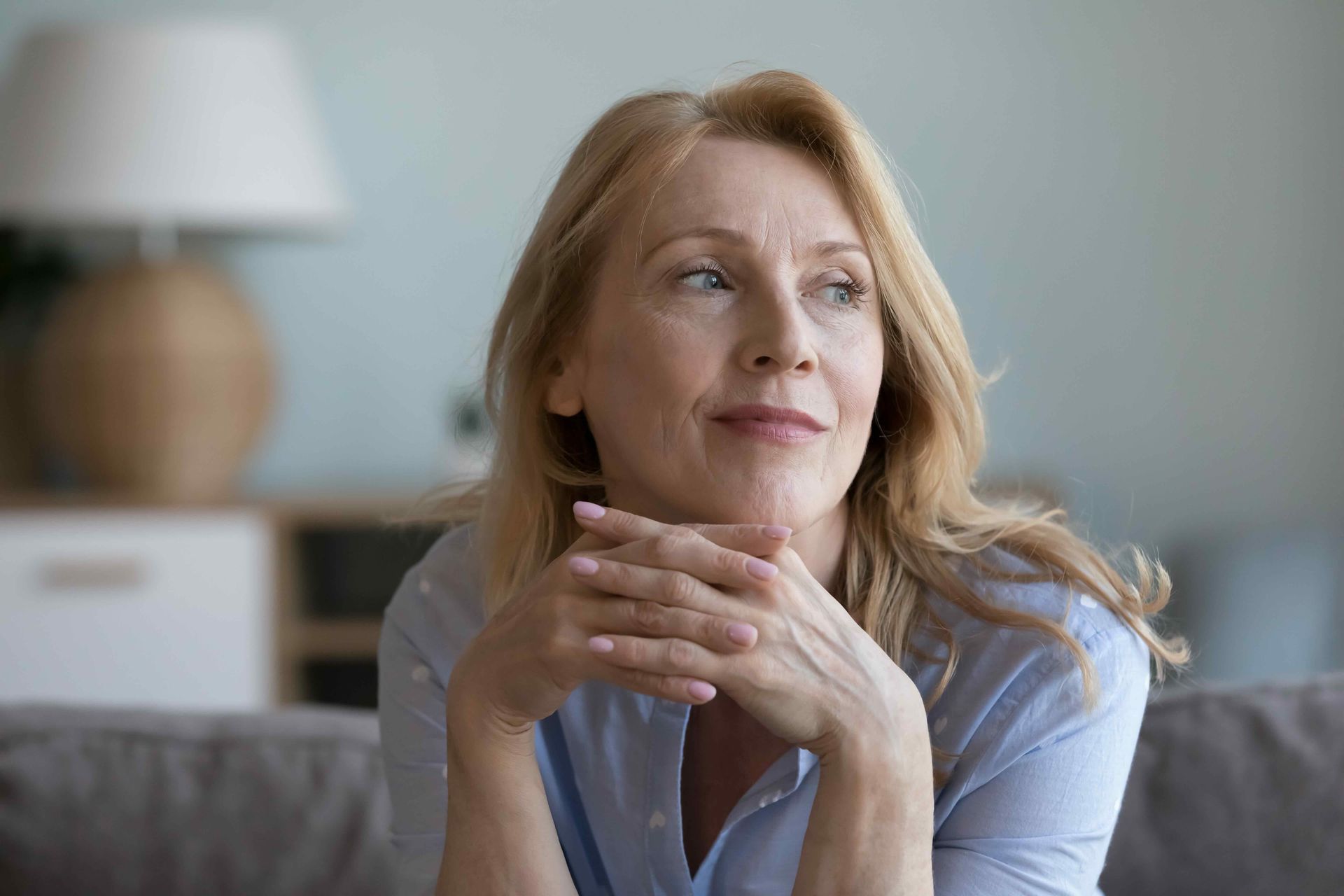 Woman in a light blue shirt resting her chin on her hands, looking off thoughtfully in a living room.