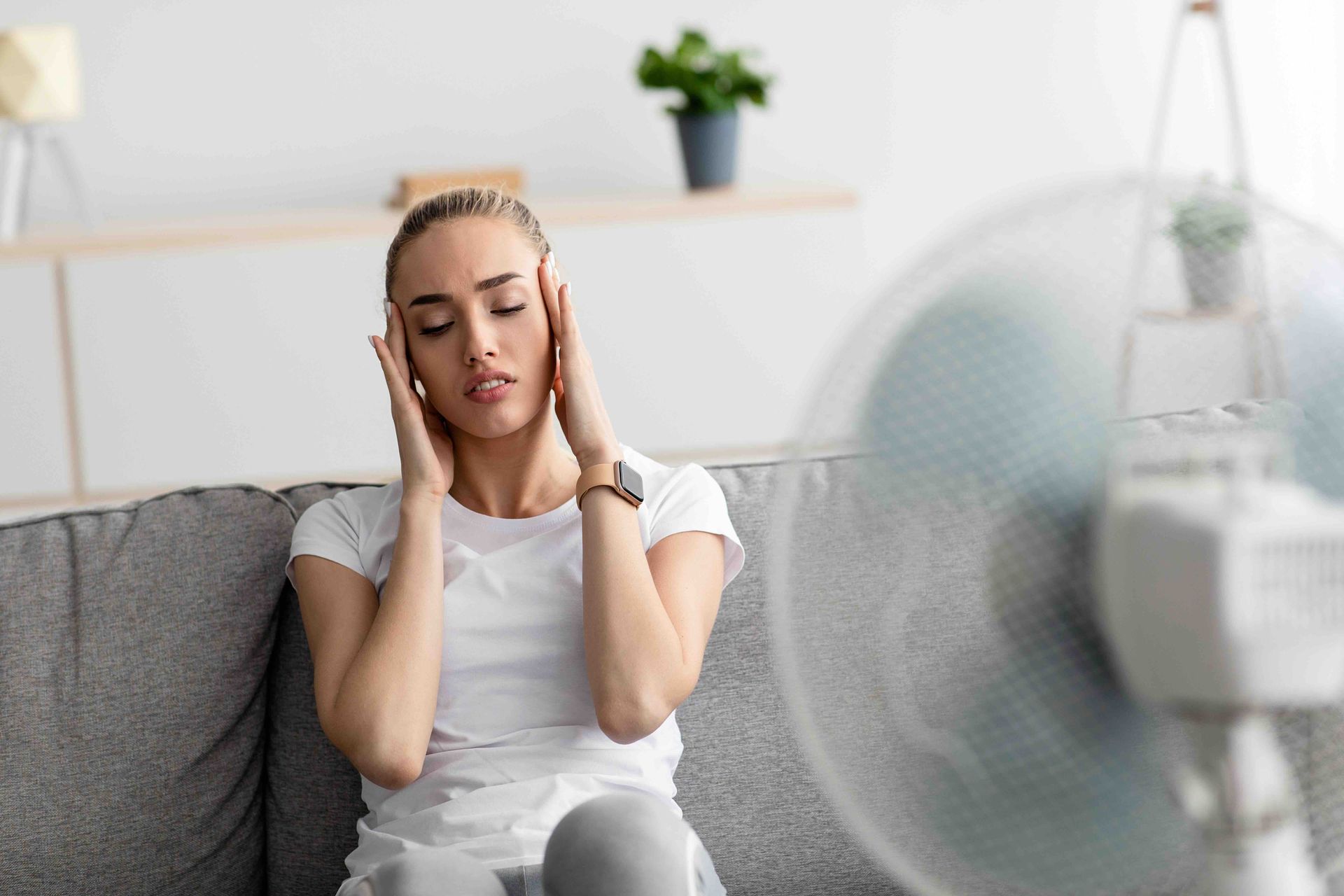 Woman with headache sits near fan on couch, indoors.