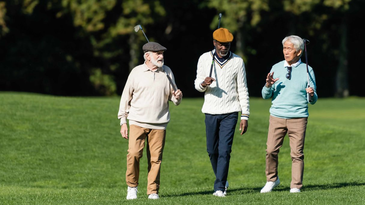 Three people walking and chatting on a sunny grassy field, each holding golf clubs