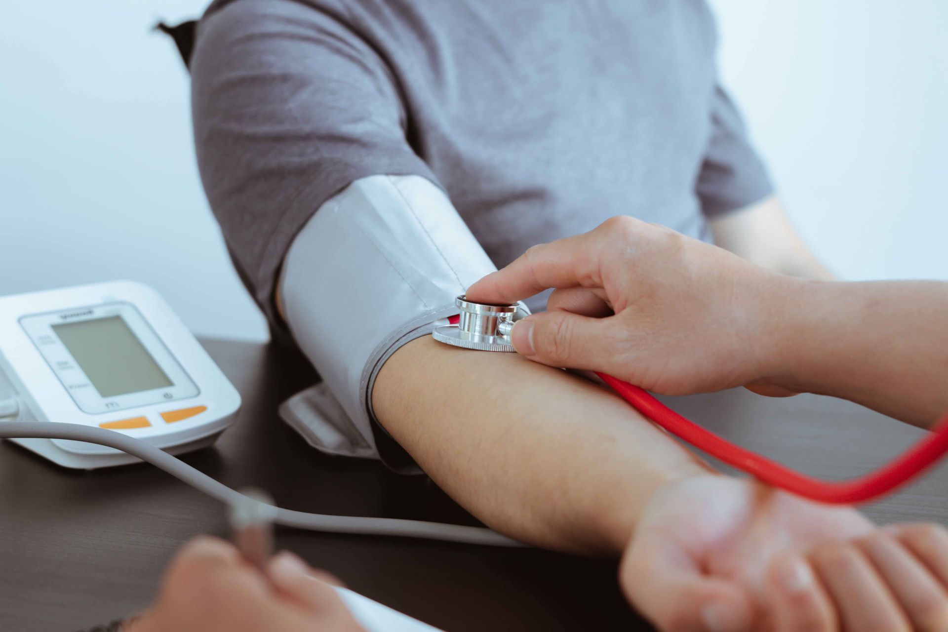 Person's arm with blood pressure cuff being examined by a stethoscope. A digital blood pressure monitor is visible.
