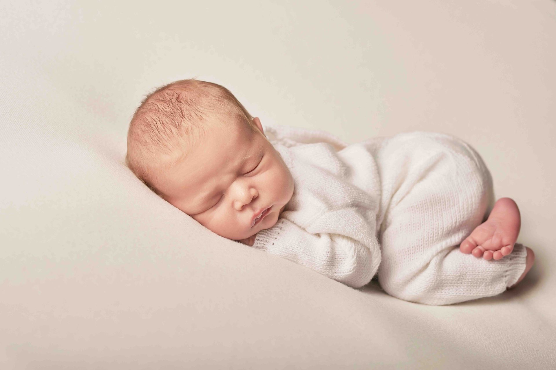 A newborn baby in white clothes sleeps curled up on a soft, light-colored background.