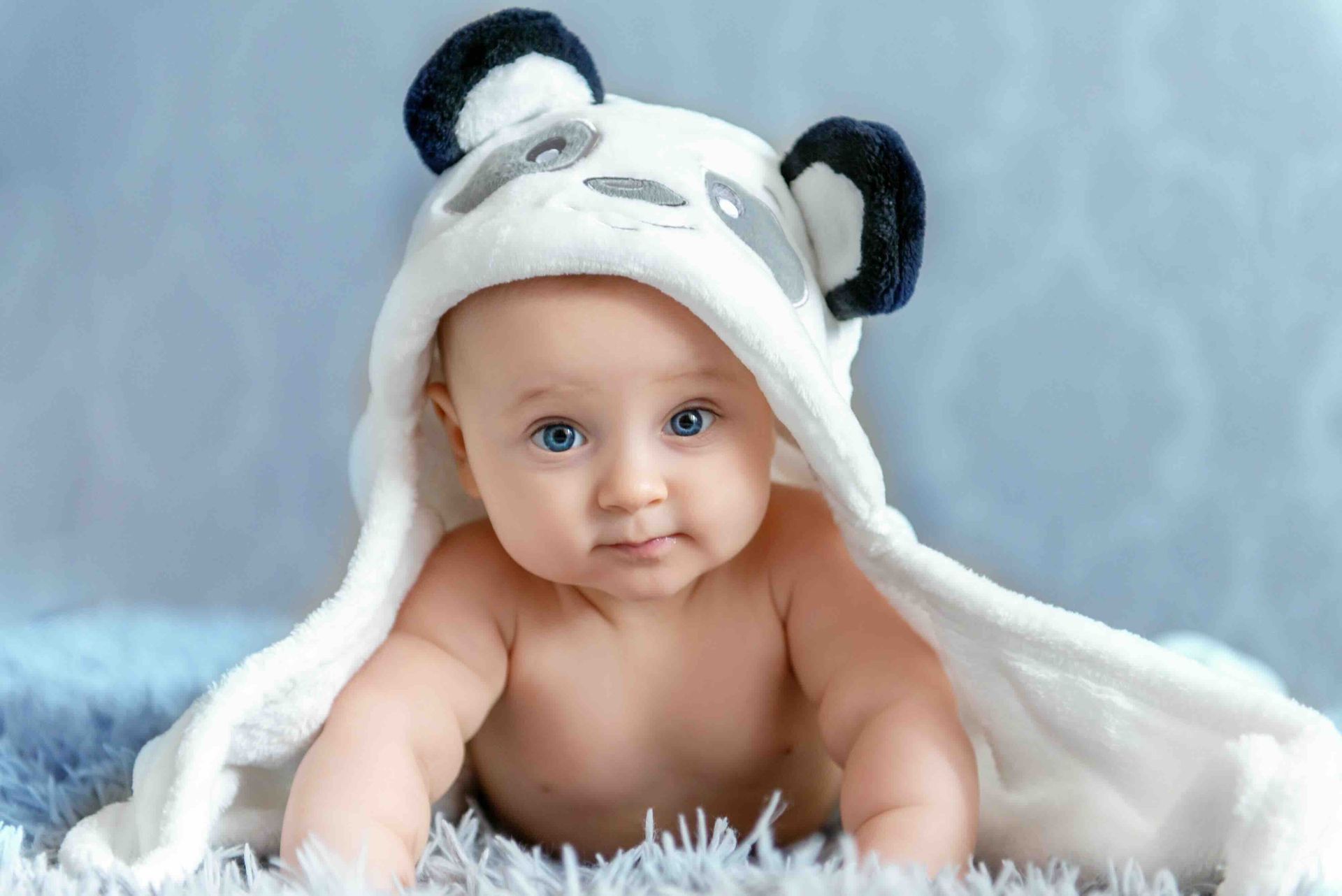 A baby wearing a hooded white and black panda towel, lying on a soft blue surface, looking toward the camera.