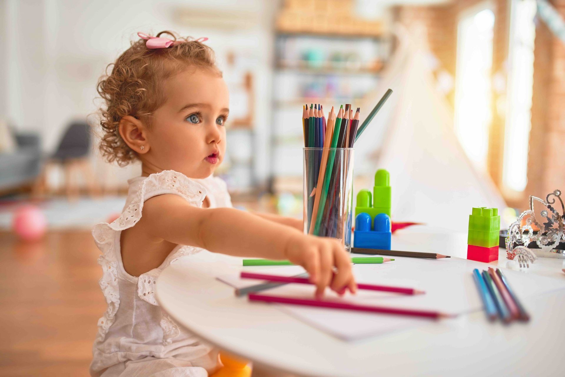 A young child with curly hair reaching for colored pencils on a white table in a sunlit room.