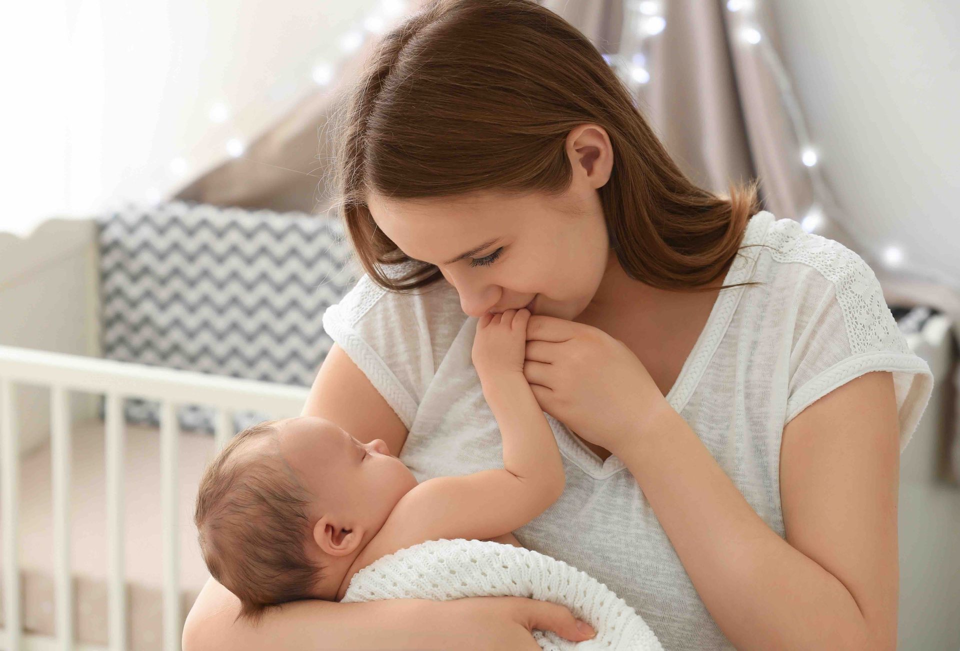 A woman is holding a newborn baby in her arms with a crib behind her.