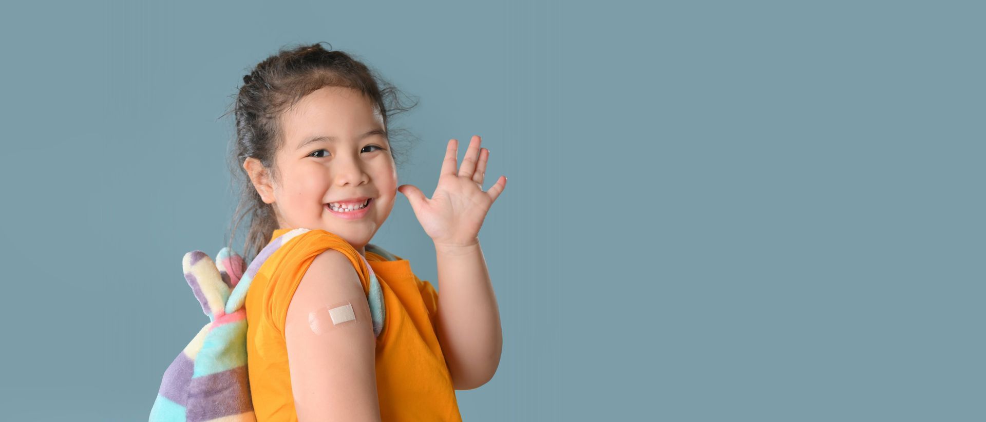 A child with a bandage on their arm waves at the camera with a smile.