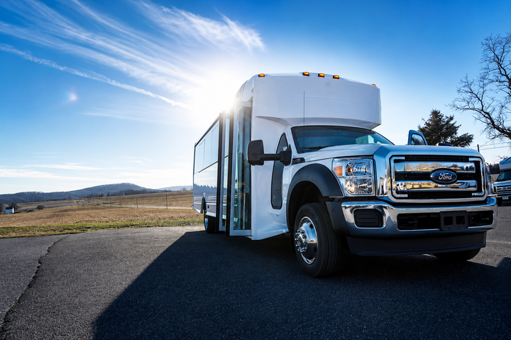 White shuttle bus on a road under a bright blue sky, with sun flare and open countryside in the background