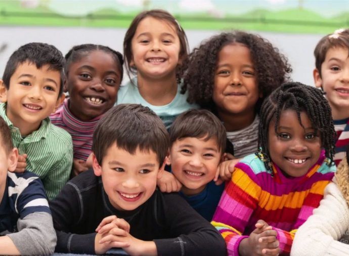 Smiling children sitting closely together indoors, wearing colorful sweaters and facing the camera.