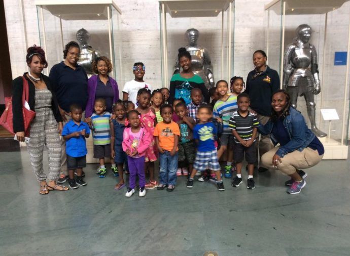 Group of children and adults posing in a museum gallery beside a large silver sculpture