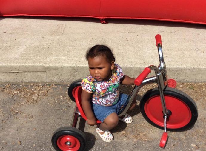 Small child on a red tricycle in a paved outdoor area, with a red curb behind.