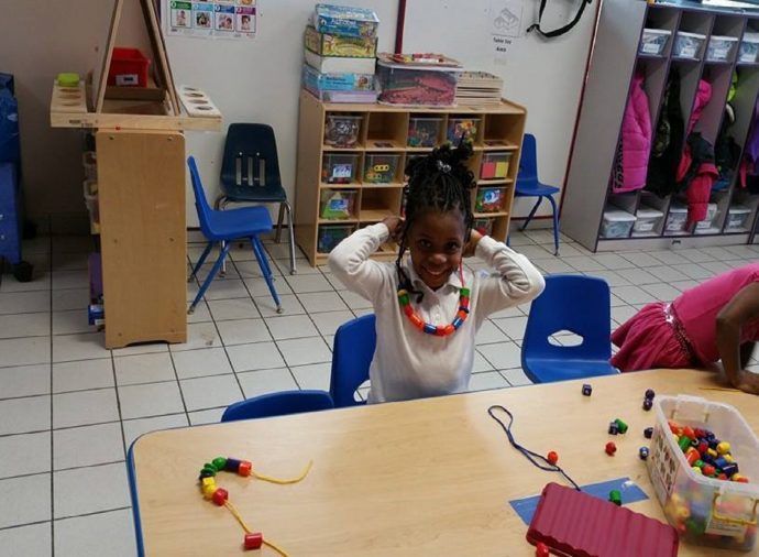 Child sitting at a classroom table with colorful beads and toys, hands on head, in a play area