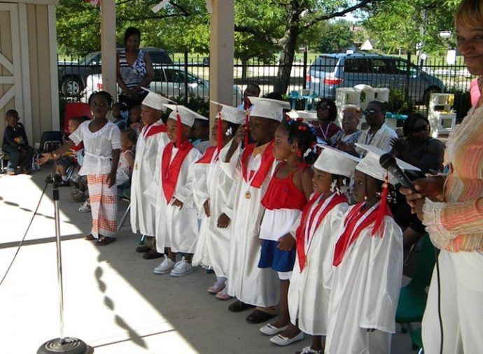 Children in white graduation gowns and red sashes singing outdoors at a ceremony