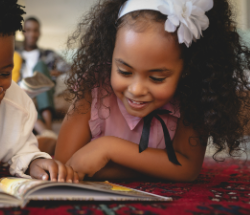 Smiling child with curly hair reading a book on the floor, wearing a pink top and white hair bow.