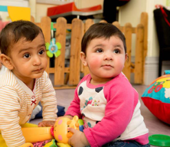 Two toddlers sitting on a playroom floor with toys, one in a pink shirt and one in a striped top