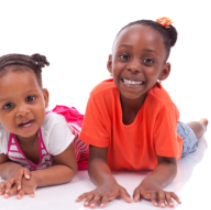Two children lying on a white floor, smiling at the camera, one in orange and one in pink.