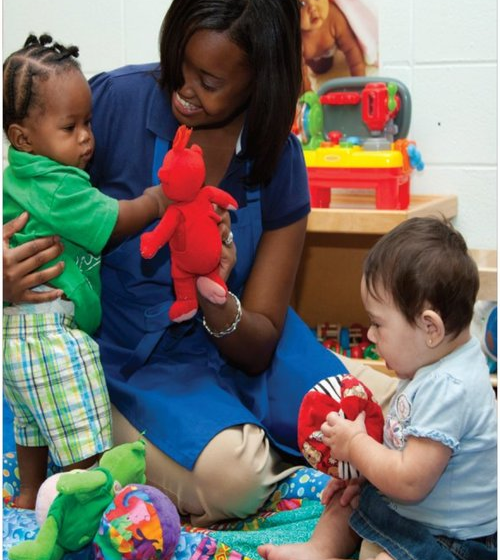 Caregiver seated with two toddlers playing with stuffed toys on a classroom mat