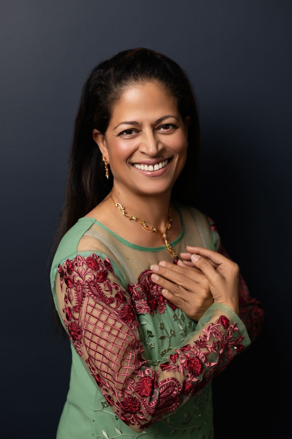 Woman in green embroidered dress smiles with hands clasped; dark blue background.