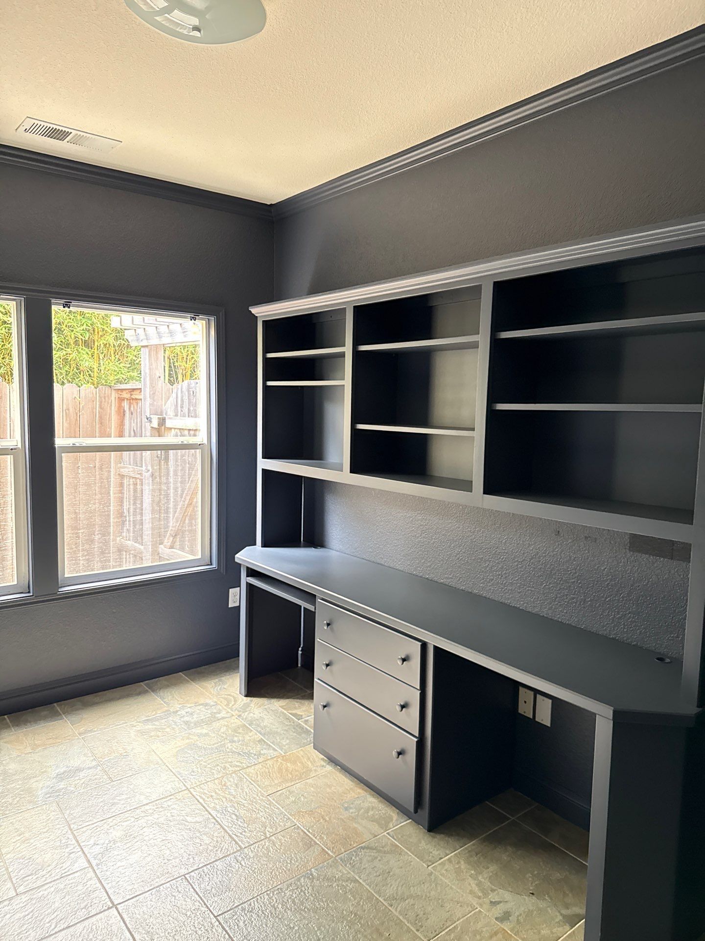 Empty home office with dark gray built-in desk, shelves, and a window on the left.