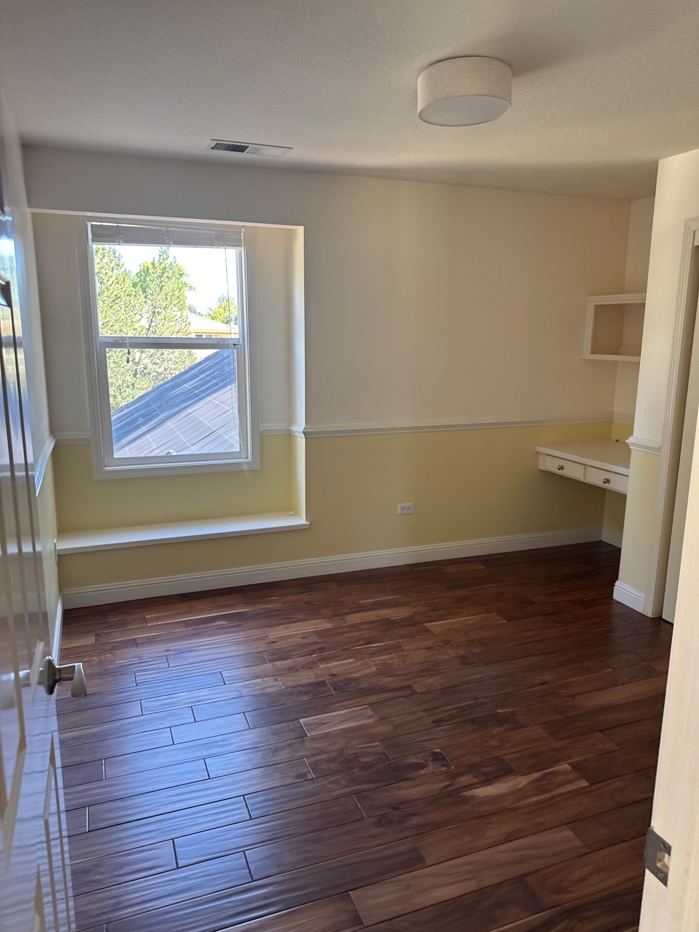 Empty beige room with wood floor, large window, and built-in shelving/desk nook.