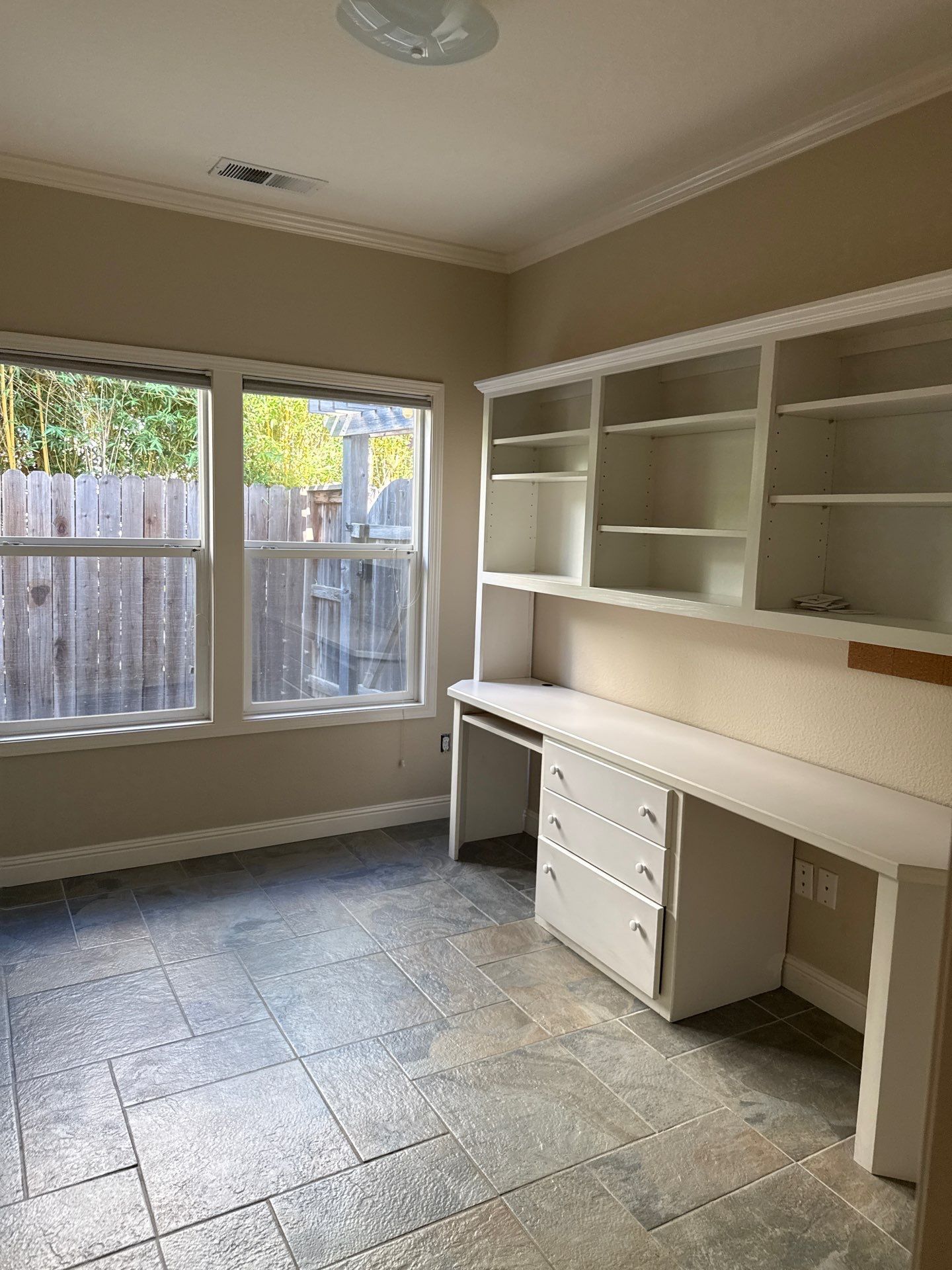 Empty room with built-in desk and shelves, tiled floor, and windows with a wood fence outside