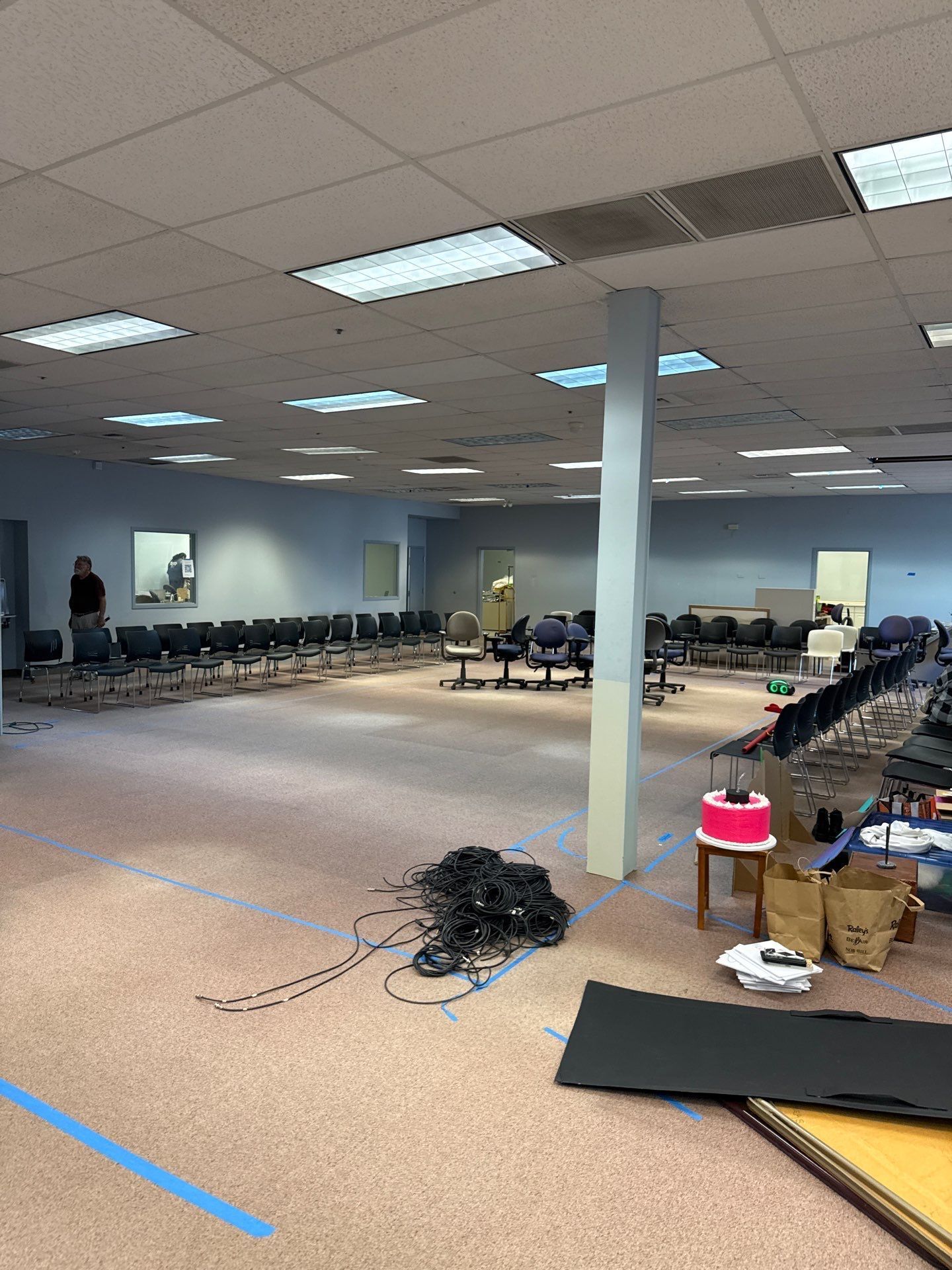 Large empty room with rows of chairs, carpeted floor, ceiling lights, and scattered cables and mats.