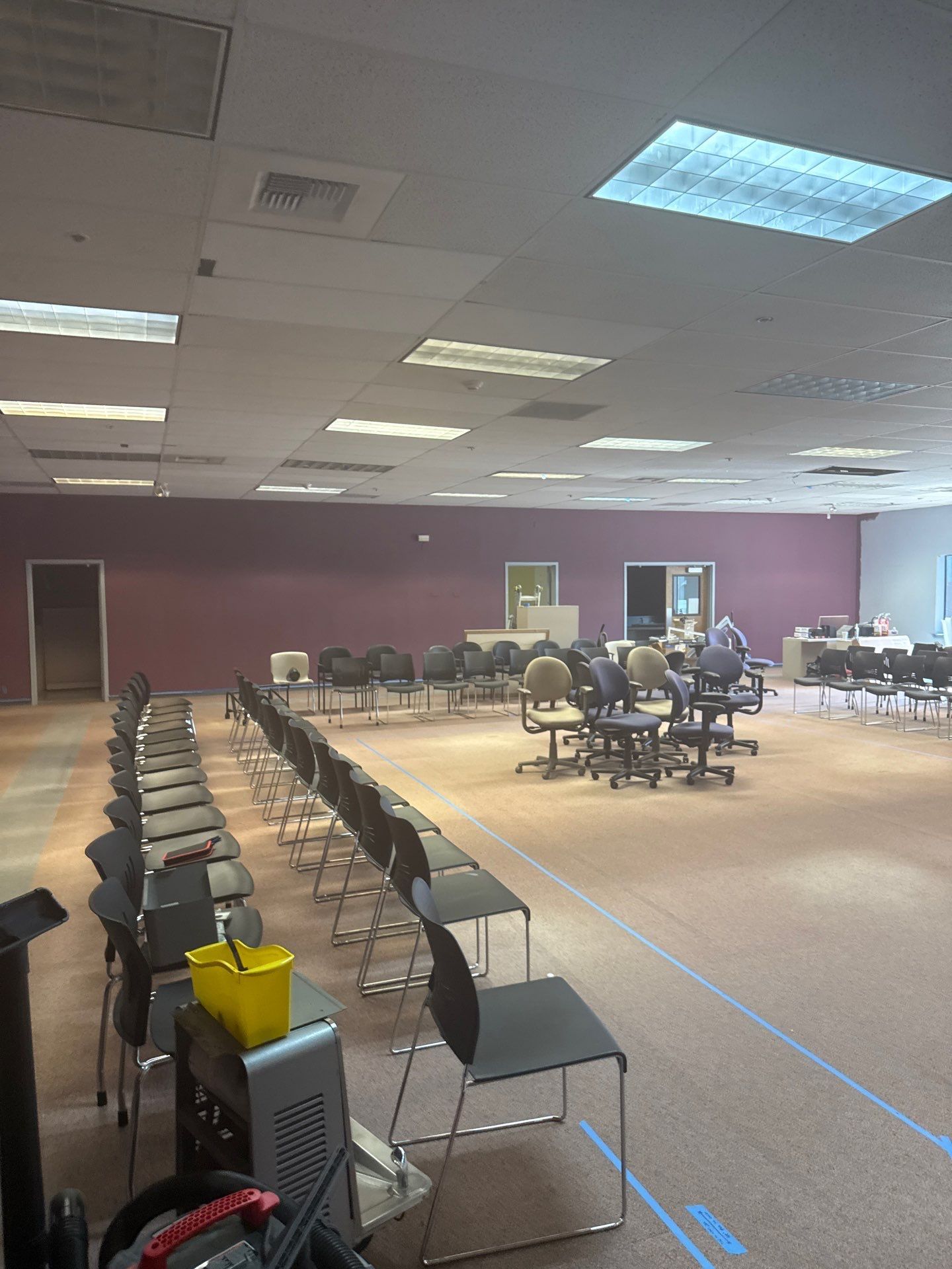 Large meeting room with rows of chairs and round tables, blue tape on the floor, and maroon walls.