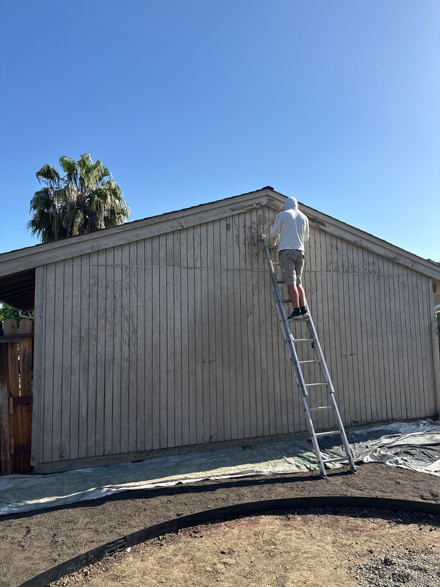 Worker on a ladder painting the side of a beige building under a clear blue sky