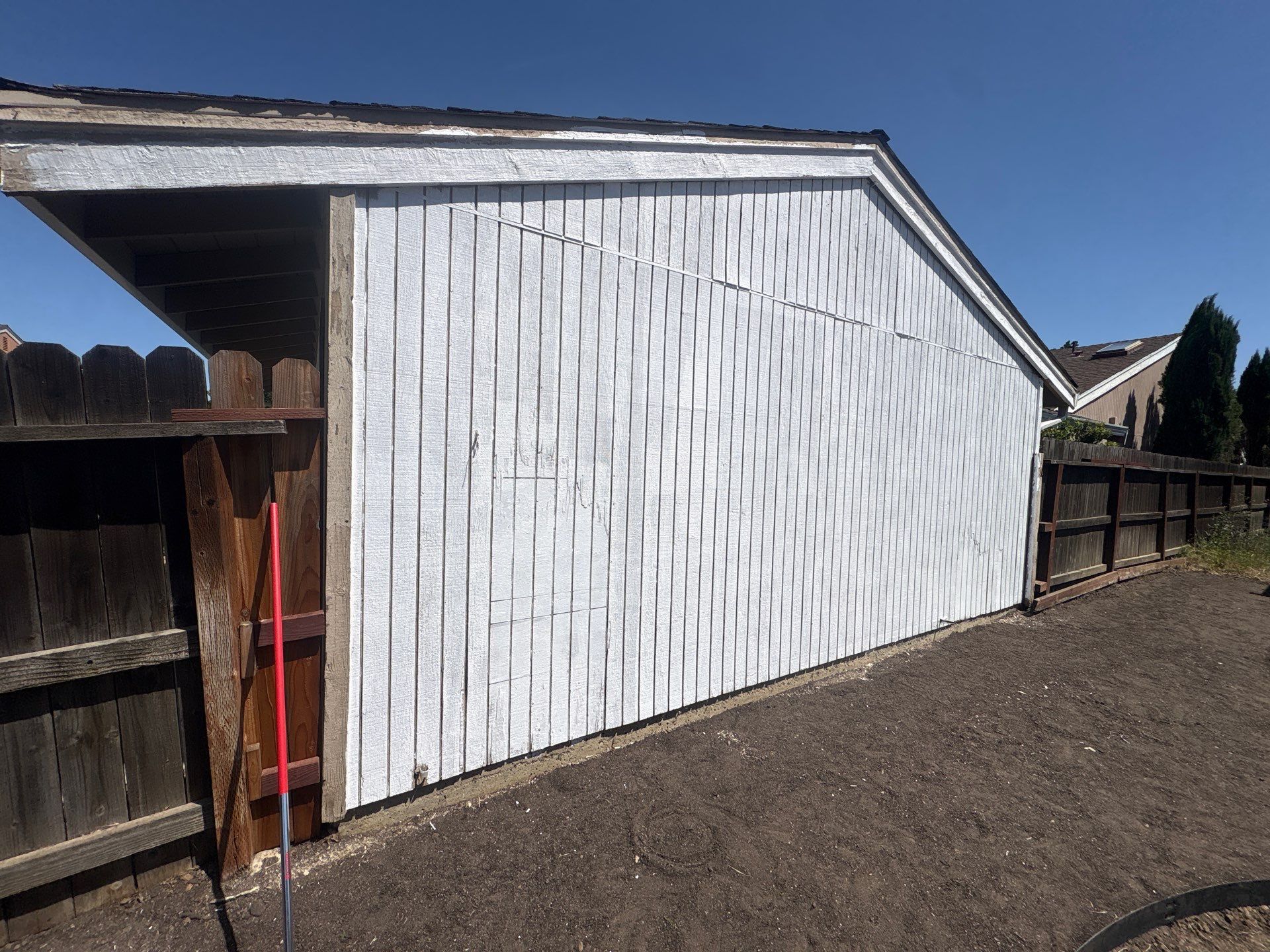 White barn or shed wall with a red-handled tool leaning near a fenced dirt yard under a blue sky
