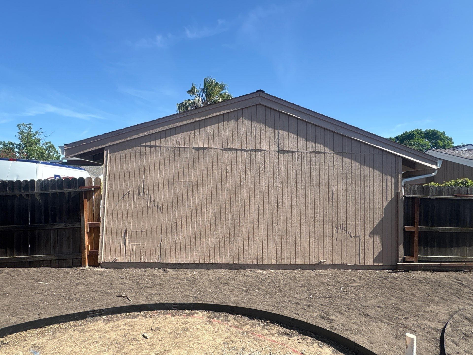 Tan-sided shed with a gabled roof in a dirt yard under a clear blue sky