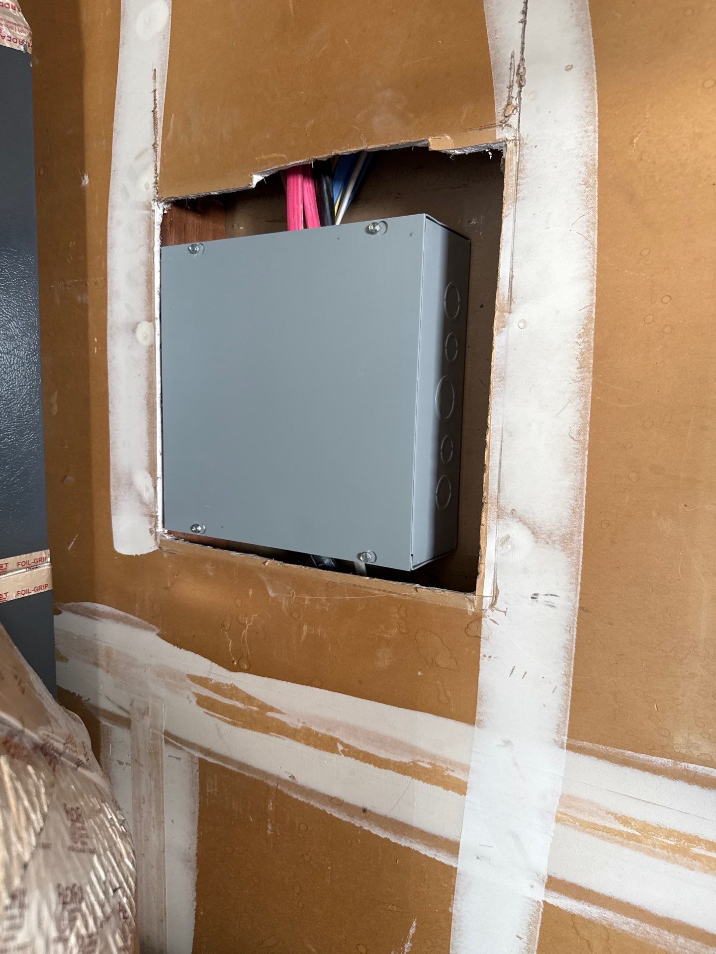 Gray electrical box mounted in a cardboard cutout, surrounded by white sealing tape