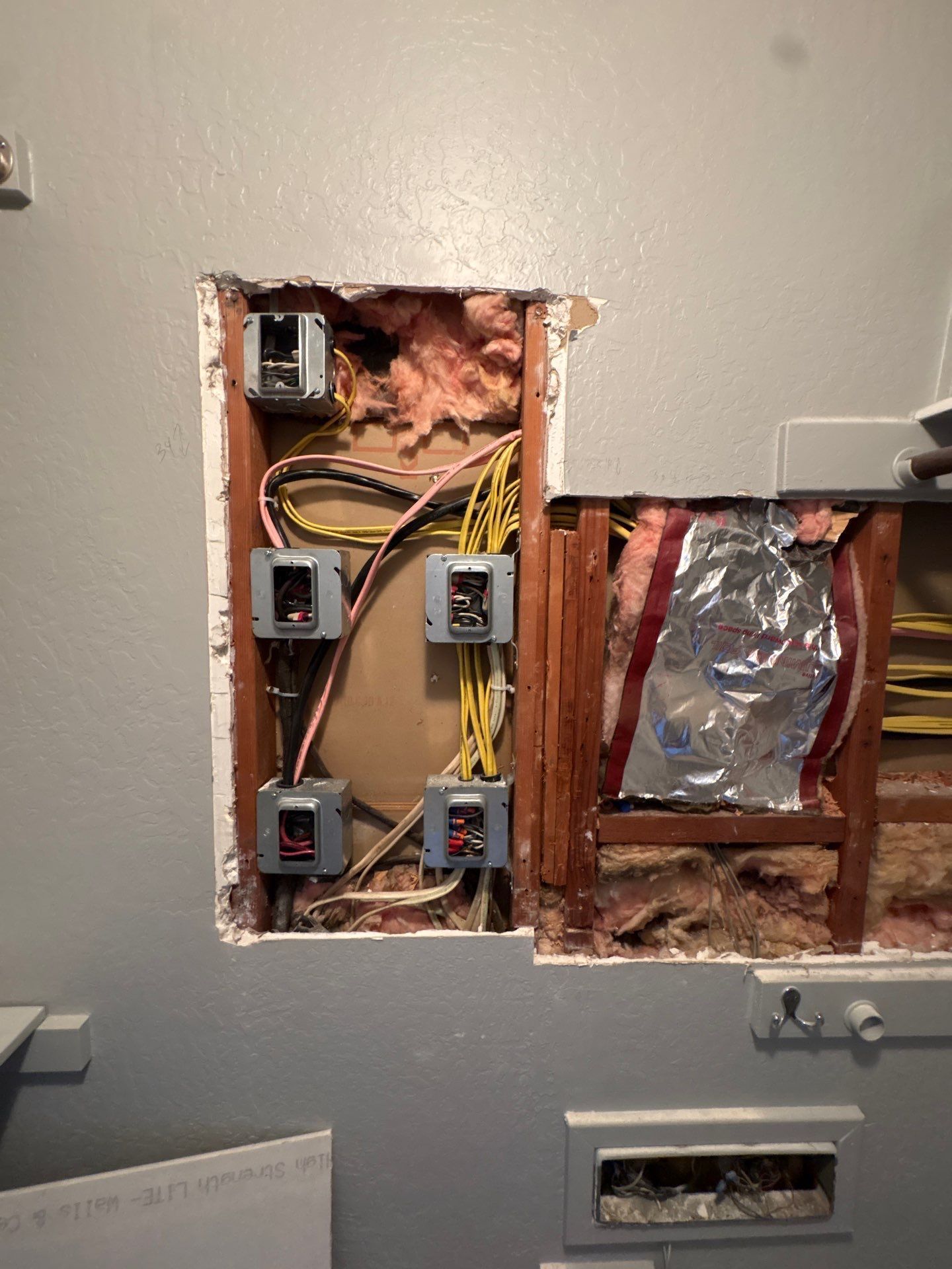 Exposed electrical wiring and boxes in a wall cavity, with foil insulation and an open outlet below.