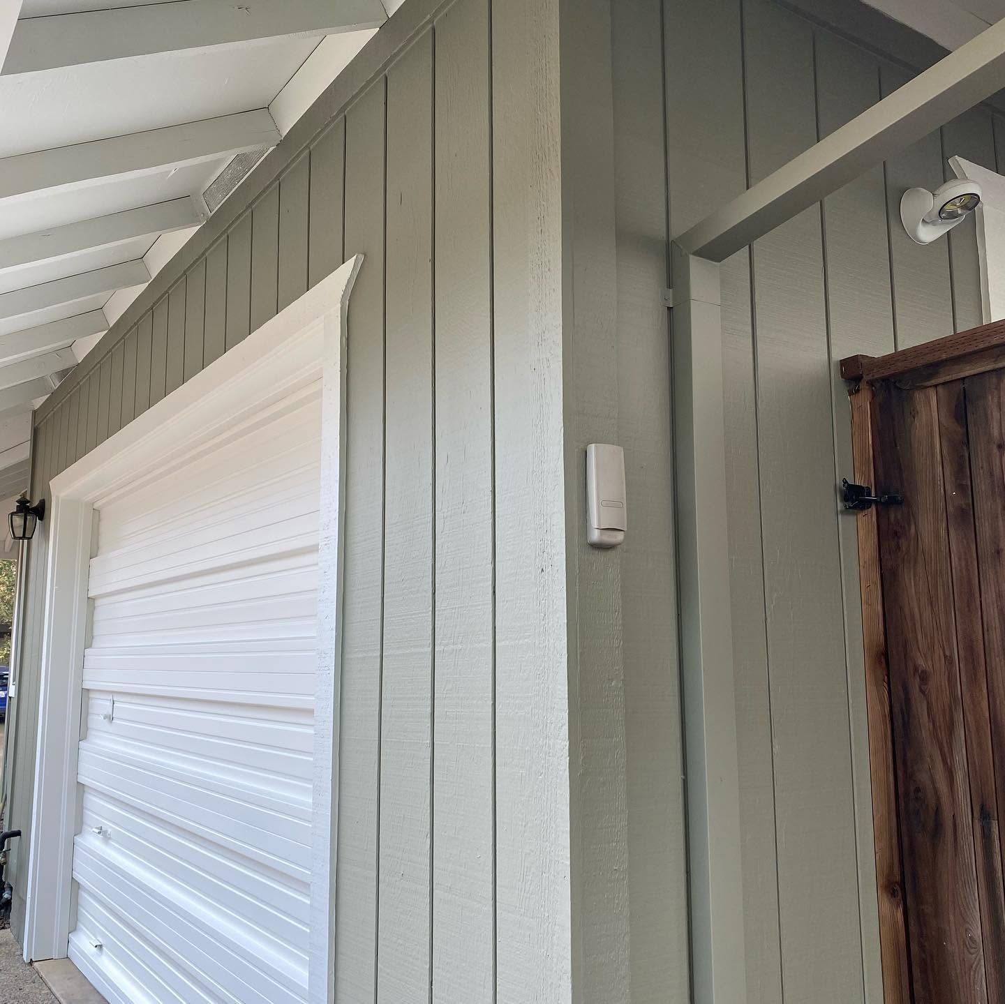 Covered exterior wall with gray siding, white garage door, and small mounted fixture by a wooden door