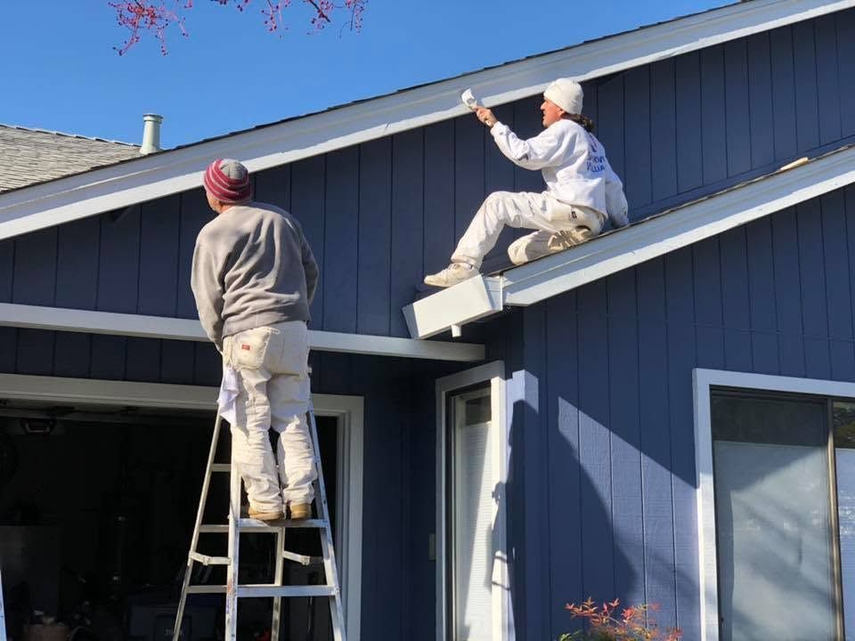Two workers paint a blue house exterior, one on a ladder and one on the roof