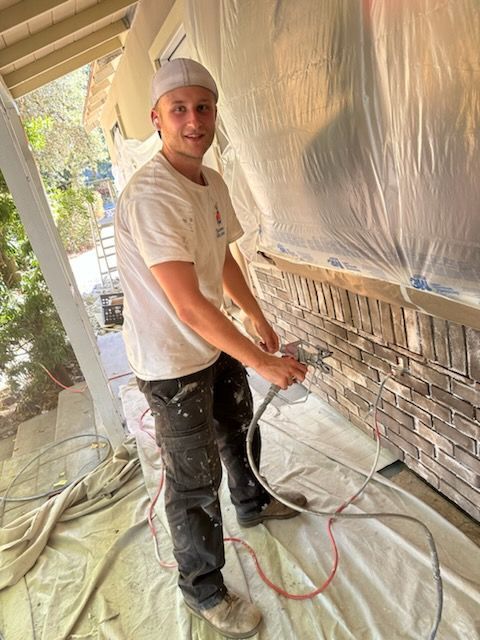 Painter spraying paint on a wall beside a stone accent panel, with plastic covering the room.