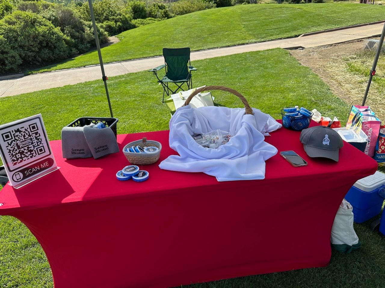 Outdoor table with red cloth, basket display, QR code sign, and small items on a grassy park lawn