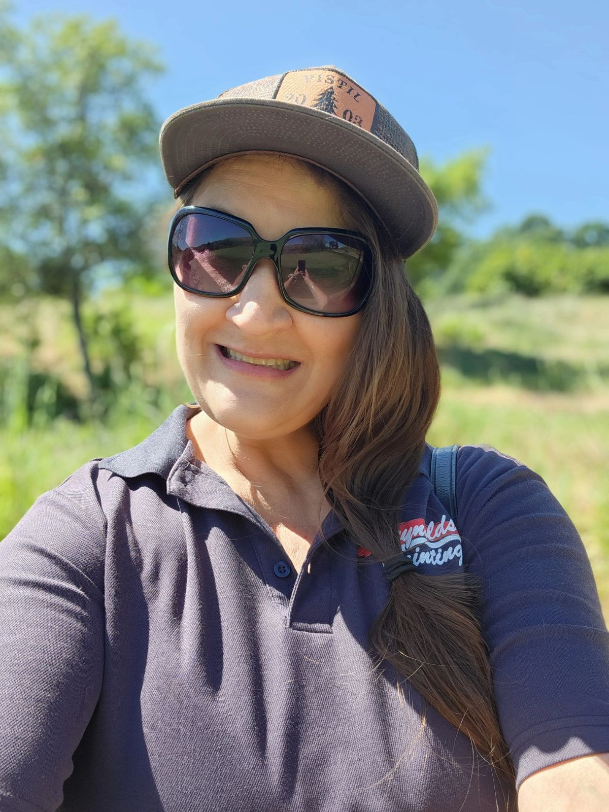 Woman wearing sunglasses and a cap, smiling outdoors in a sunlit green field.