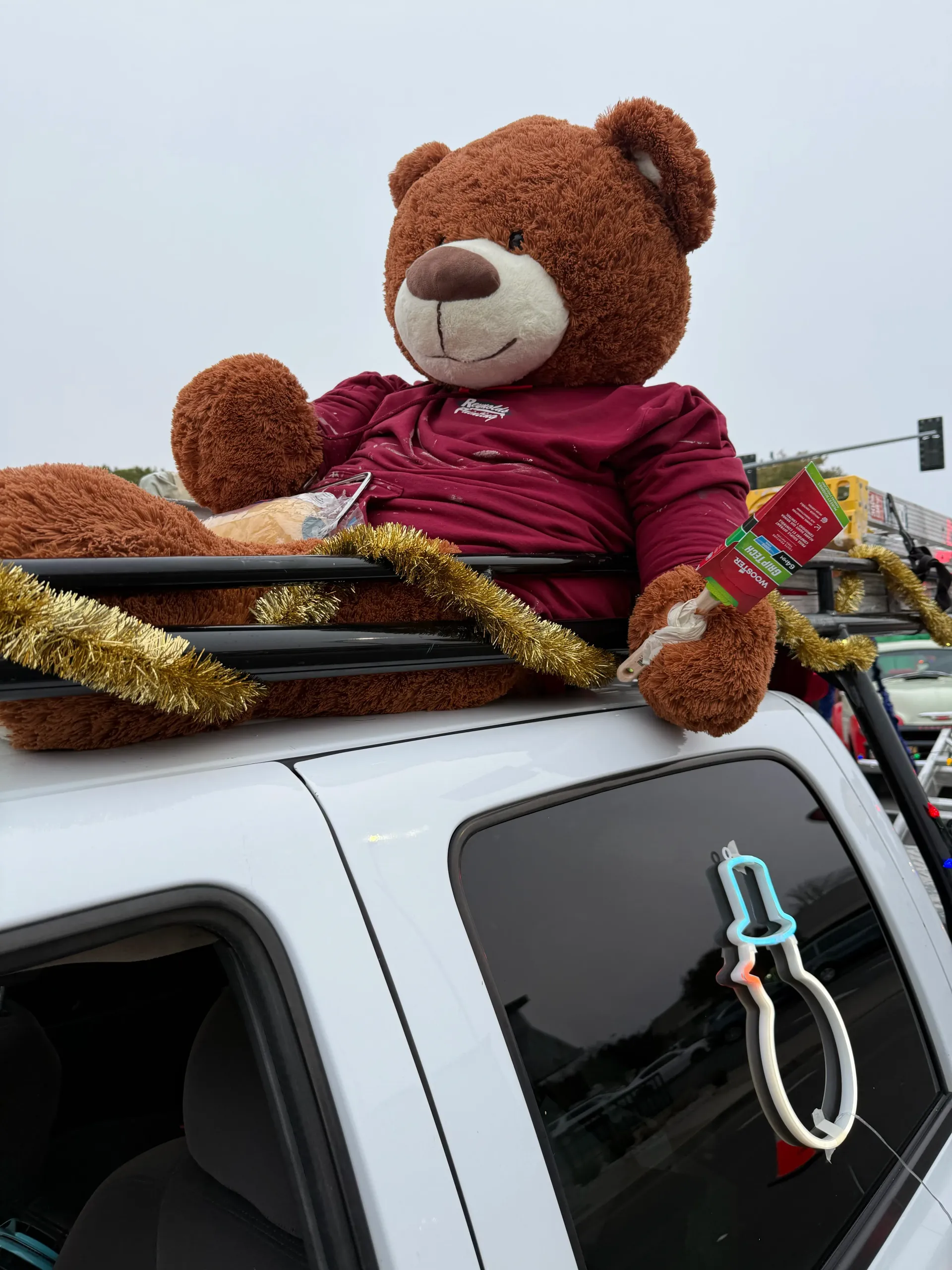 Large teddy bear in a maroon shirt riding atop a decorated vehicle.
