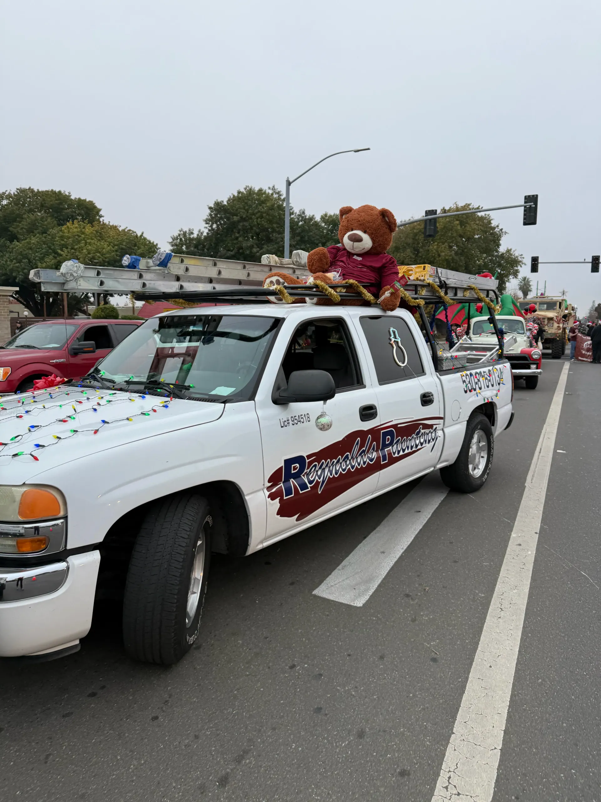 Decorated white pickup trucks in a parade on a city street, one with a teddy bear on top