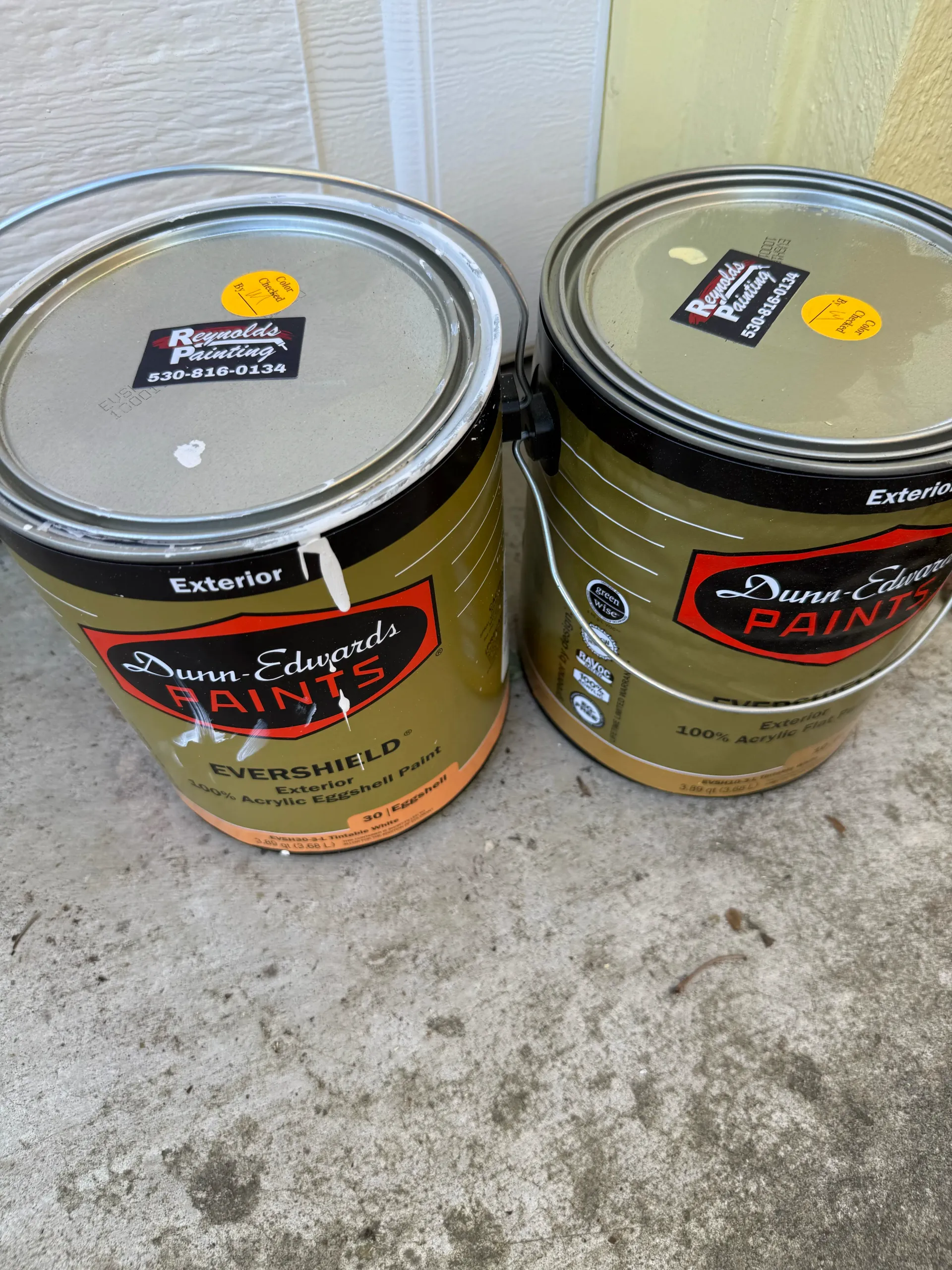 Two paint cans on a concrete floor, one gray and one olive green, each with a lid and label.