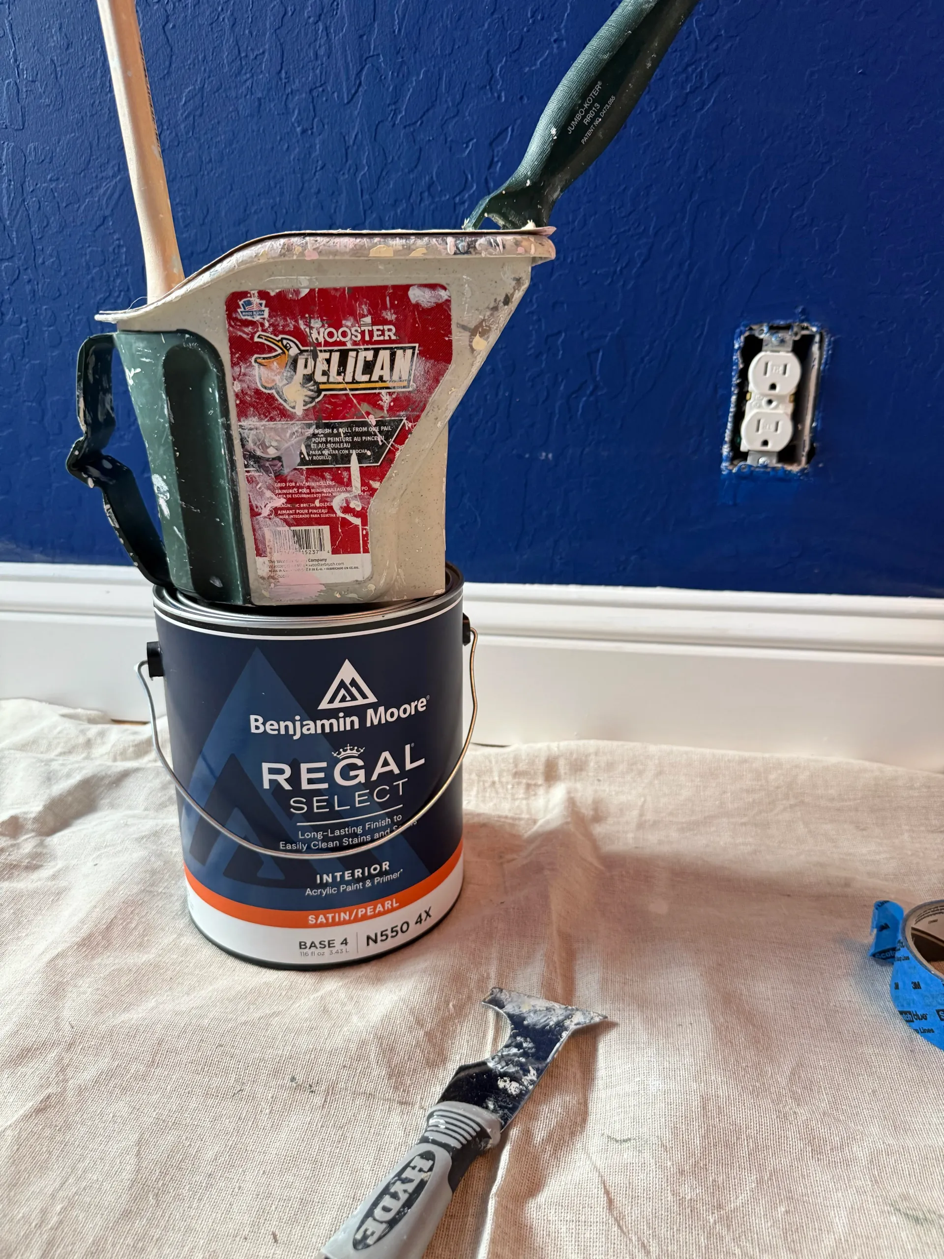 Paint scraper and putty knife standing in a paint can on a drop cloth beside a blue wall outlet.